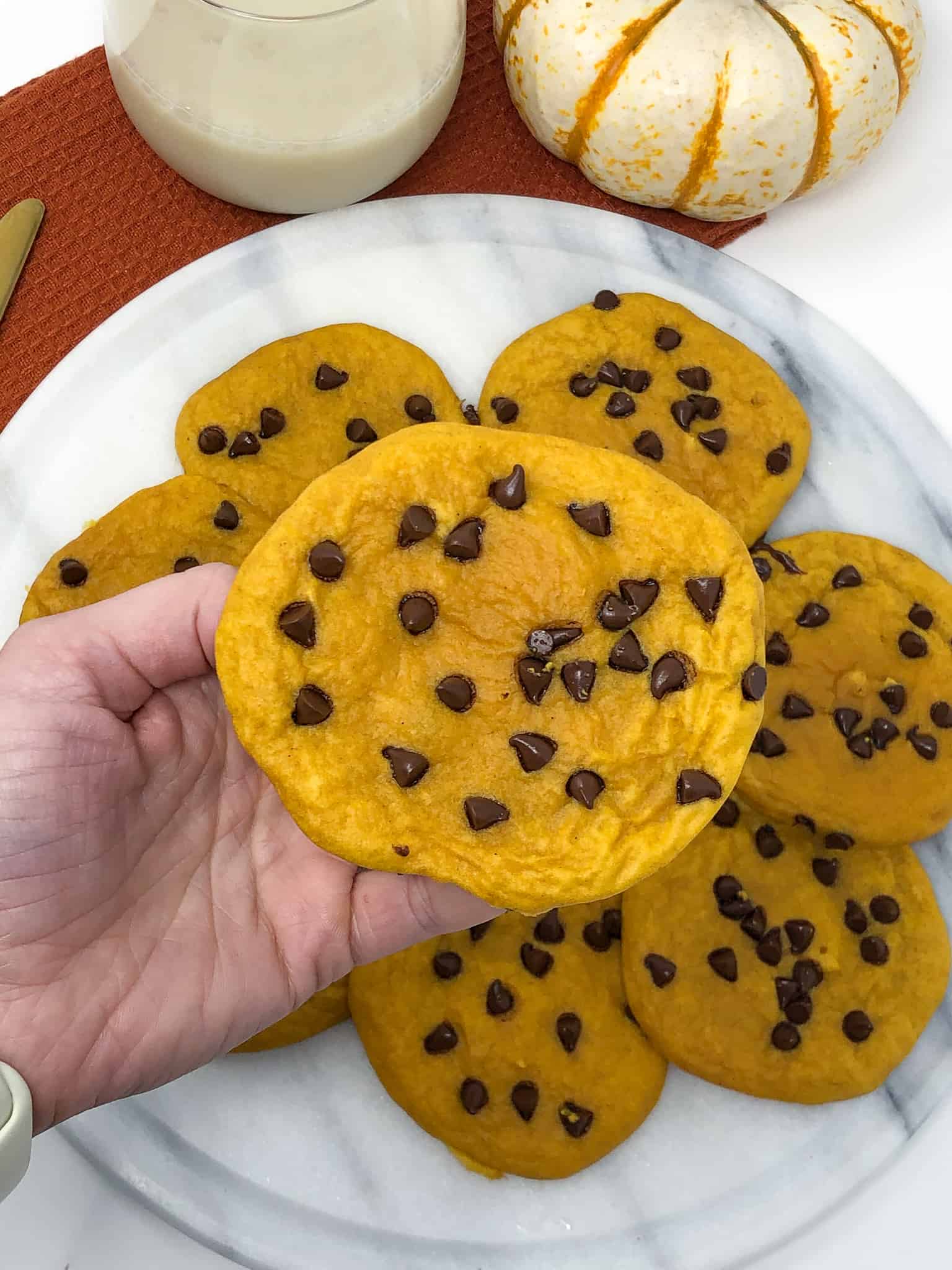 A hand holding a pumpkin chocolate chip cookie with a plate of cookie in the background.