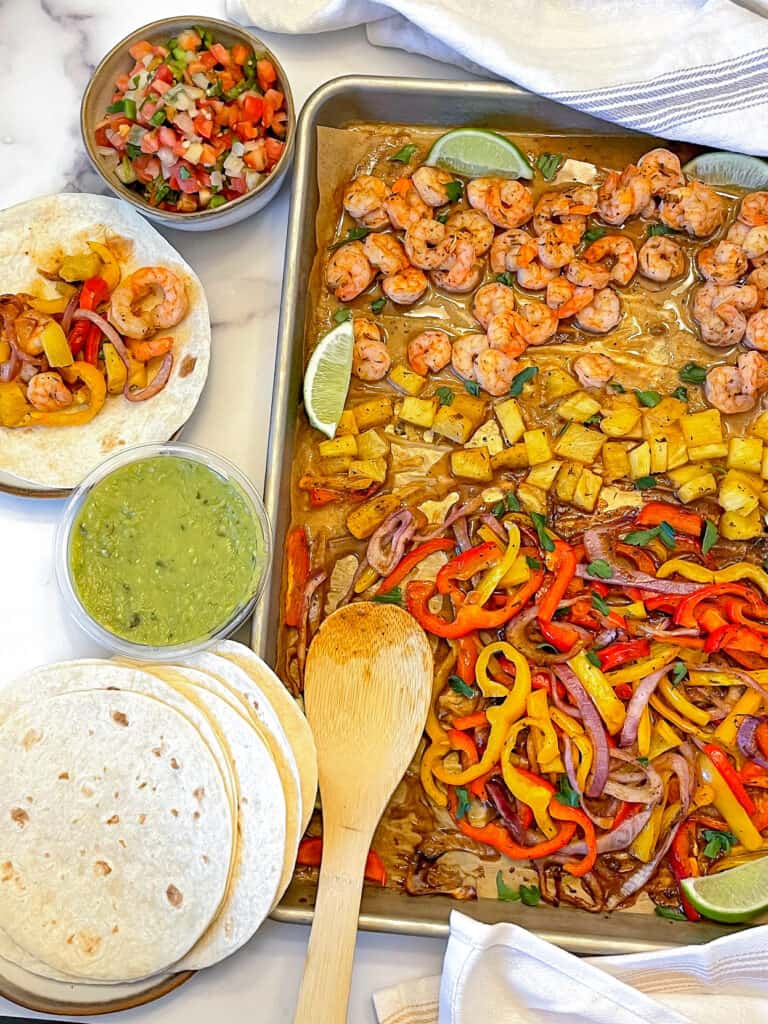 Shrimp fajitas on a sheet pan with tortillas, guacamole, and pico de gallo on the side.