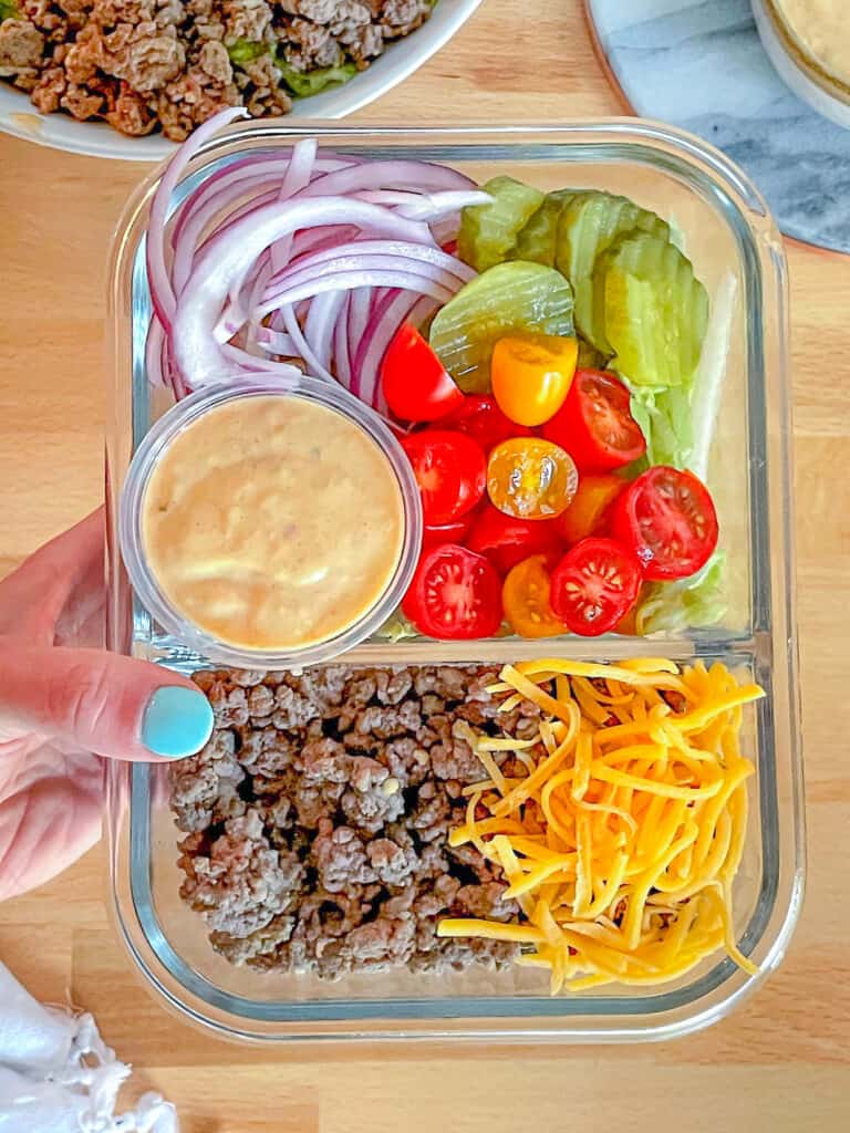 Meal prep cheeseburger bowl in a meal prep container with a hand holding a side of skinny special sauce.