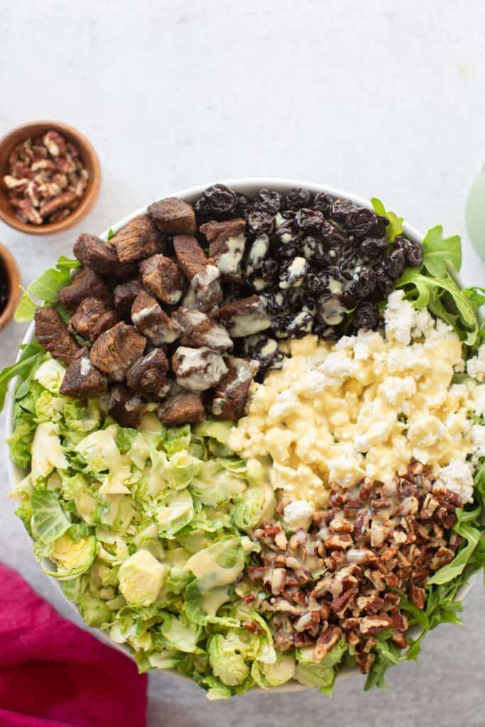 A brussels sprouts salad with steak bites, pecans, feta cheese, dried cranberries, and dressing in a large bowl.