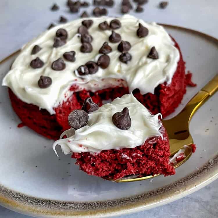Red velvet mug cake topped with frosting and chocolate chips on a plate with a fork.