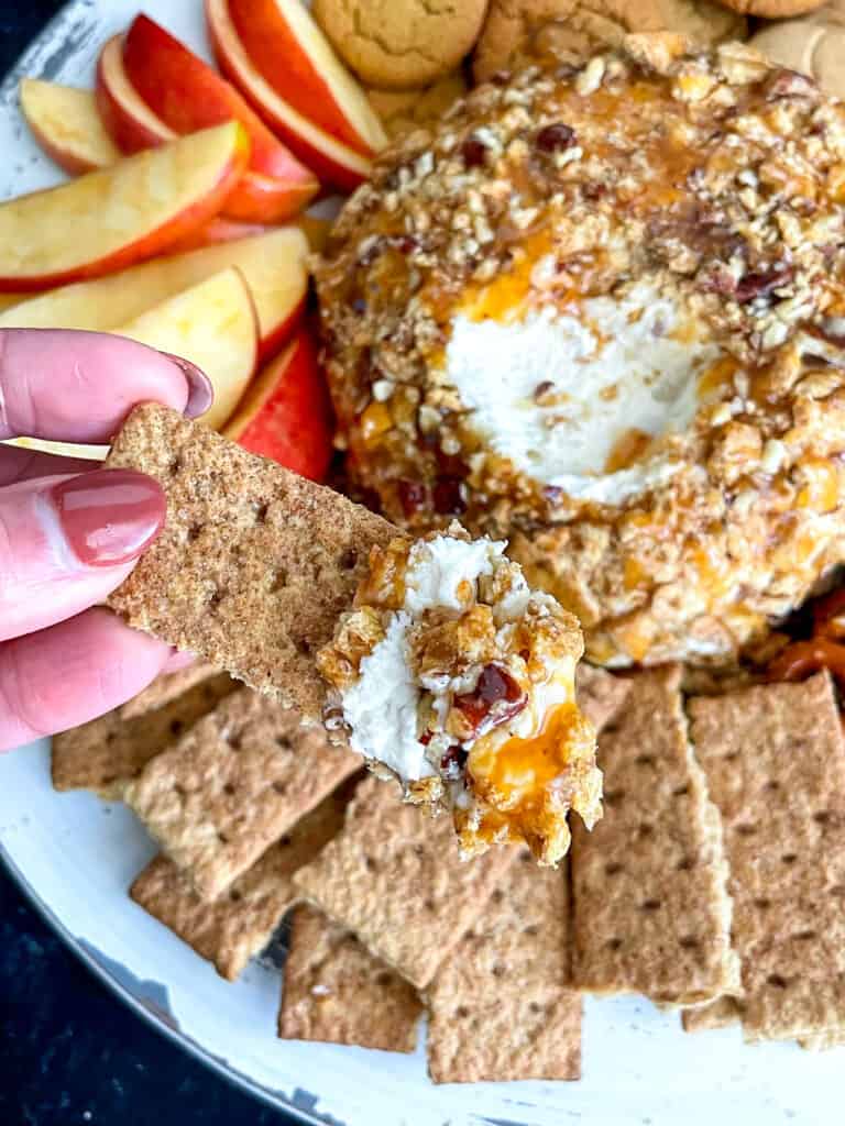 A hand dipping a graham cracker into caramel pecan dessert cheese ball with addtional graham crackers, cookies, and apple slices on the side.
