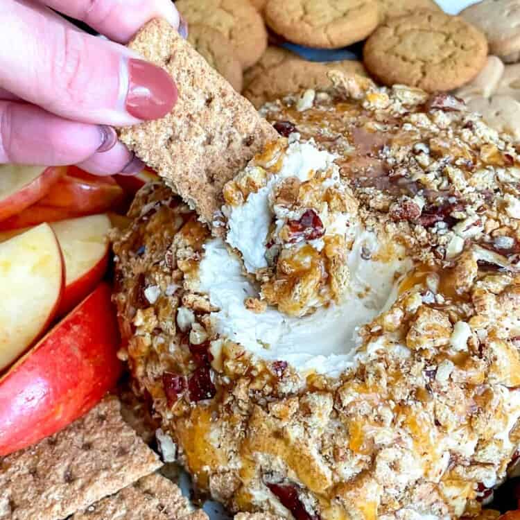 A hand dipping a graham cracker into caramel pecan dessert cheese ball with addtional graham crackers on the side.