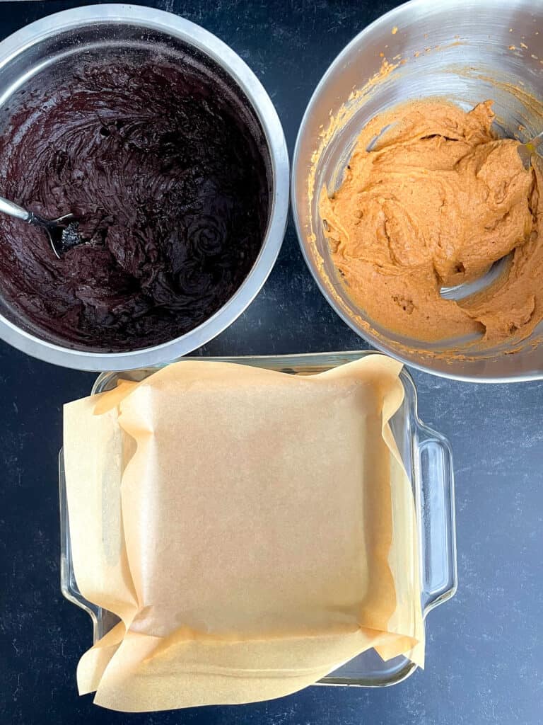 Pumpkin and brownie batter in mixing bowl and a square pan lined with parchment paper.