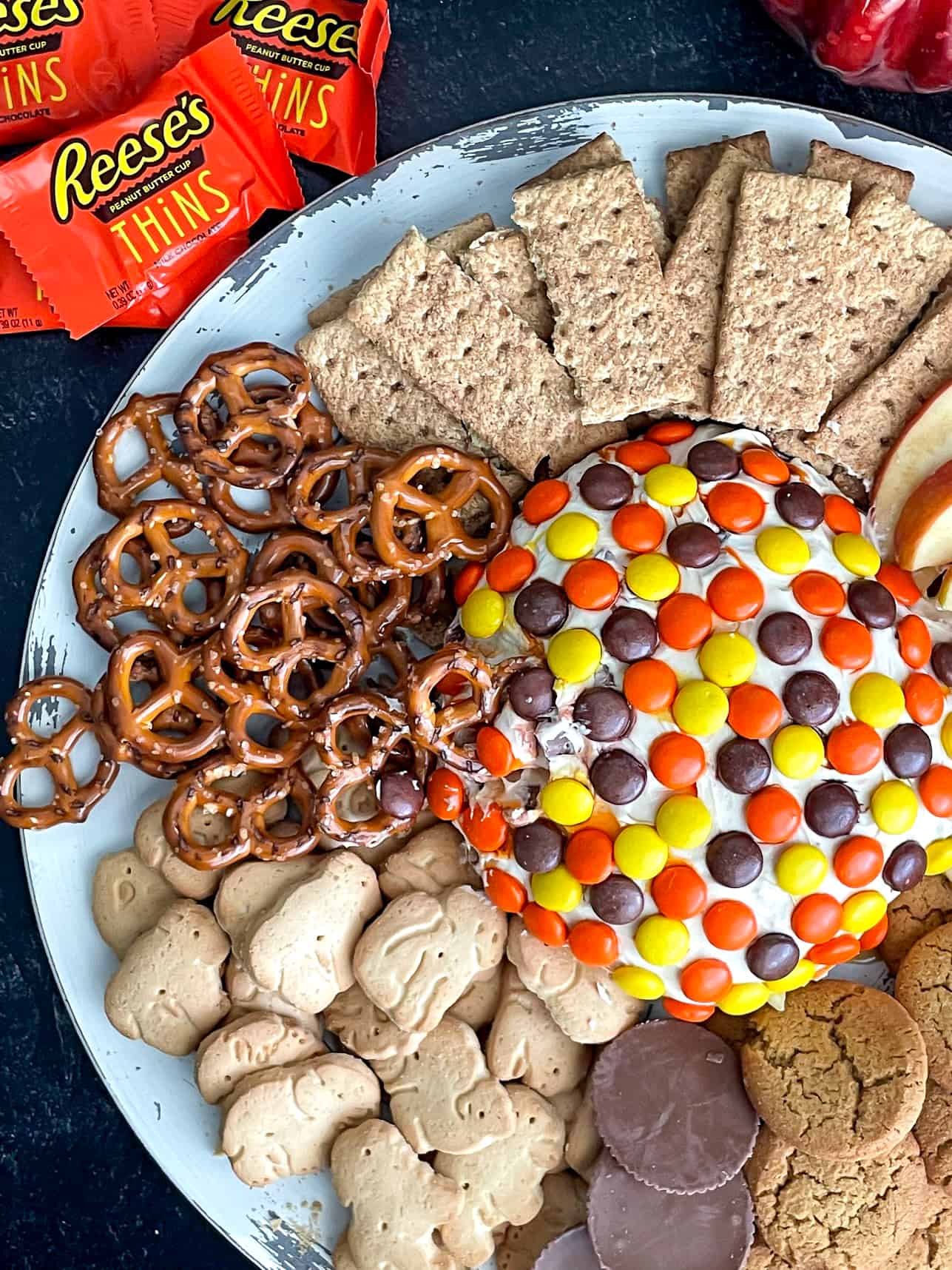 Peanut butter cheese ball topped with Reeses Pieces on a tray with pretzels, animals crackers, graham crackers, and cookies.