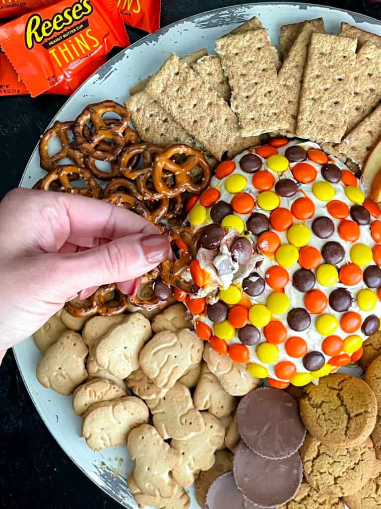 A hand dipping a pretzel in peanut butter cheese ball topped with Reeses Pieces on a tray with pretzels, animals crackers, graham crackers, and cookies.
