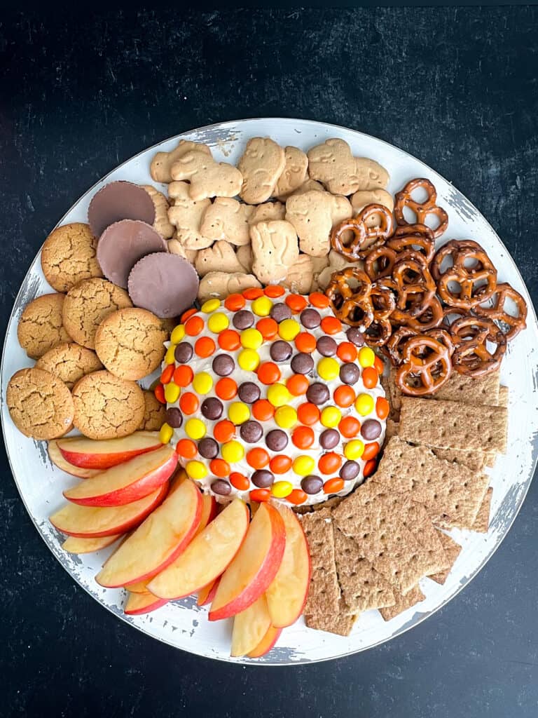 Peanut butter cheese ball topped with Reeses Pieces on a tray with pretzels, animals crackers, graham crackers, and cookies.