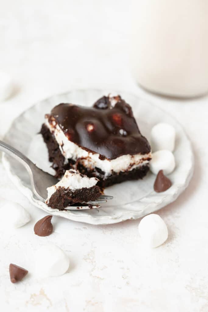 One Mississippi Mud Brownie on a white plate with a fork surrounded by marshmallows and chocolate chips.