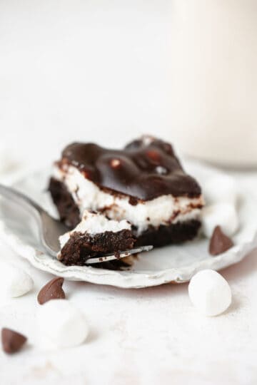One Mississippi Mud Brownie on a white plate with a fork surrounded by marshmallows and chocolate chips