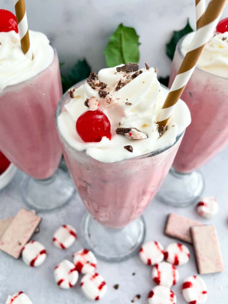 Skinny peppermint milkshakes topped with whipped cream, peppermint pieces, and a cherry in a tall glasses with straws.