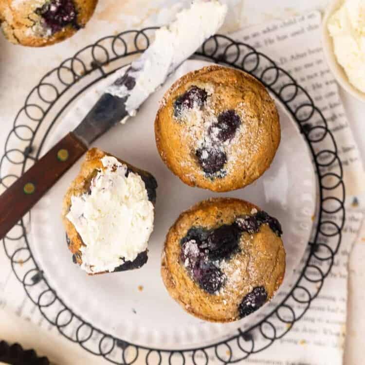 Blueberry muffins on a plate with a knife, one cut in half and spread with butter.