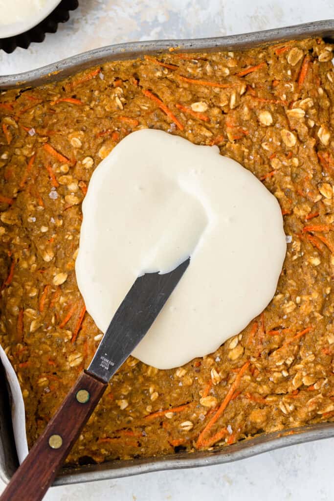 Carrot cake oatmeal bake being spread with vanilla maple cream cheese icing in a baking pan.