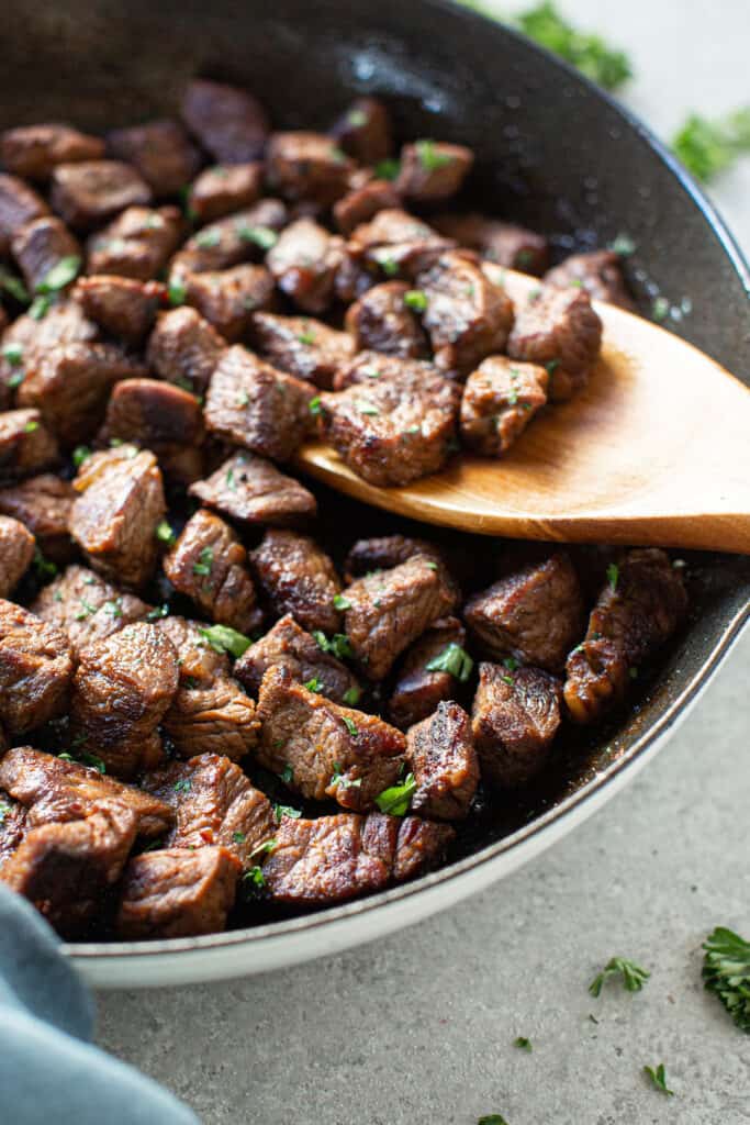 Zoomed in view of garlic steak bites in a skillet with a wooden spoon.