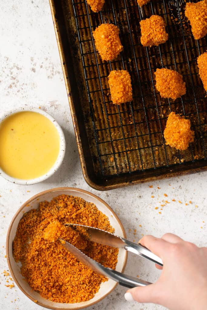 A piece of chicken being held with tongs being dipped a small bowl of breadcrumb and more coated nuggets on a cooling rack on a baking sheet.