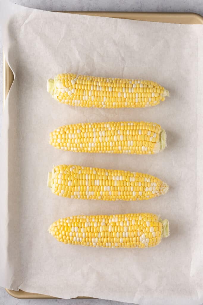 Four corn on the cobs on a baking sheet with parchment paper.