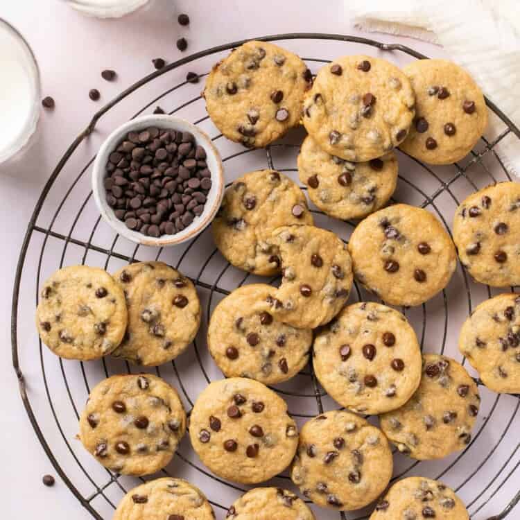 Healthy vegan chocolate chip cookies on a cooling rack alongside glasses of milk and a small bowl of chocolate chips.