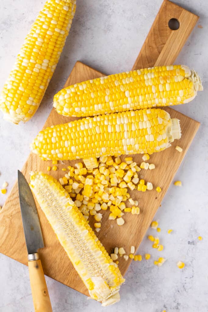 Corn being slices off the cobs with a knife on a cutting board.
