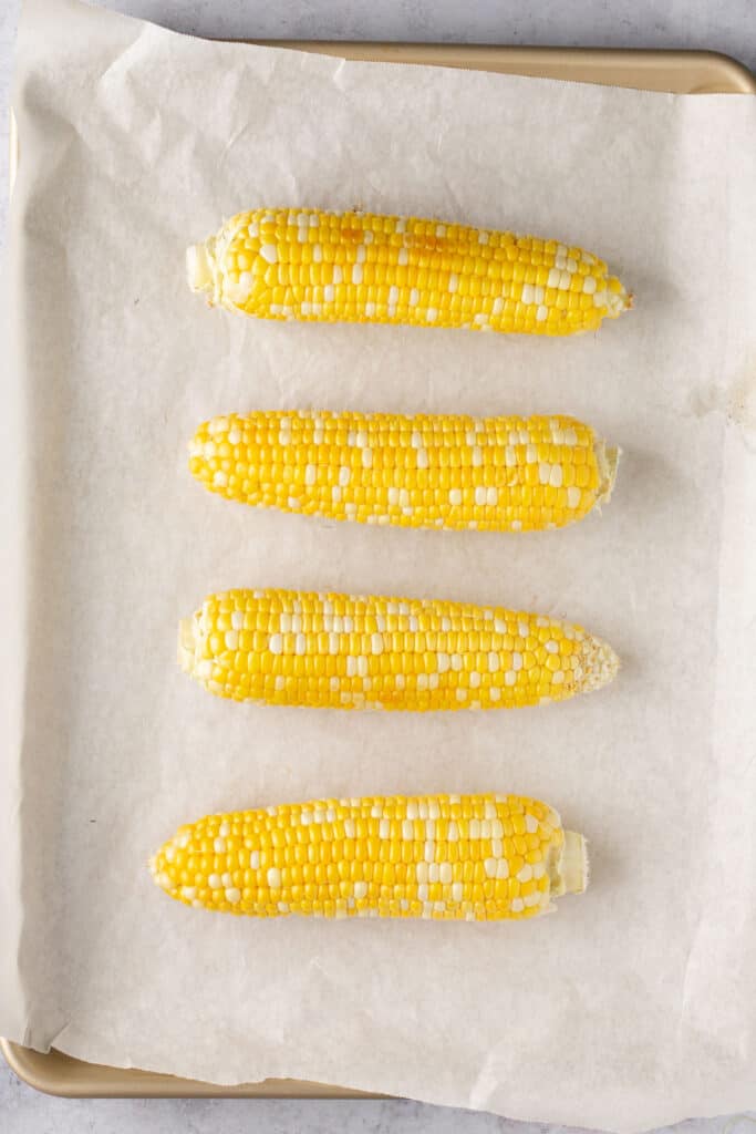 Four corn on the cobs on a baking sheet with parchment paper.