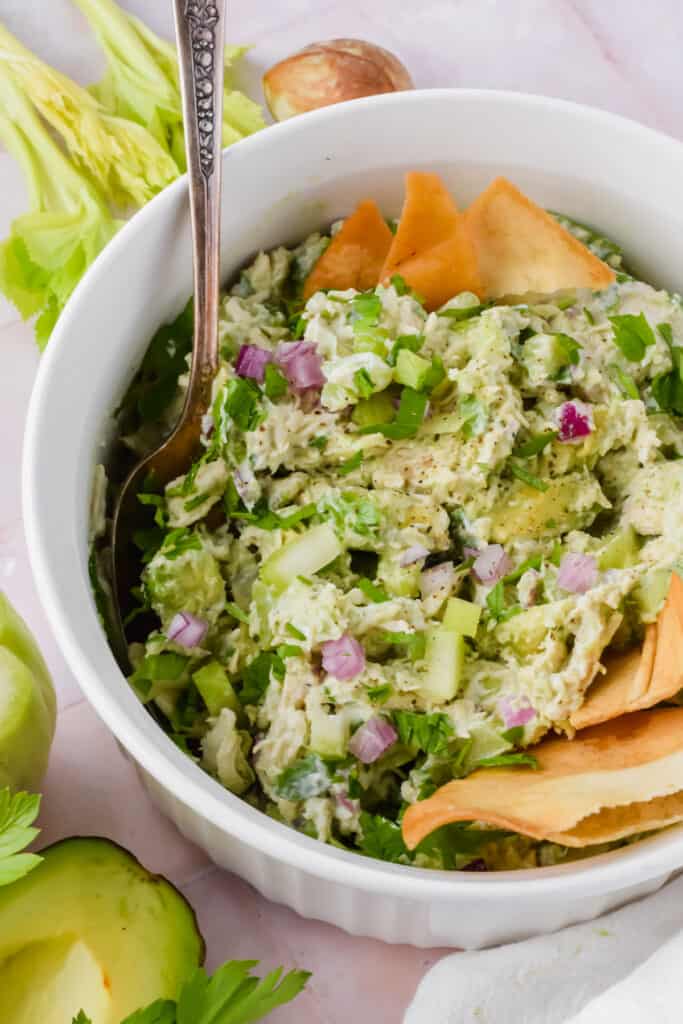 Close up view of creamy avocado greek yogurt chicken salad in a bowl with a spoon garnished with pita chips.
