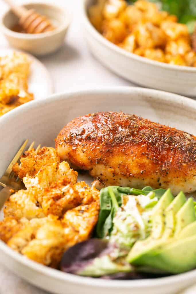 Close up view of honey garlic baked chicken and cauliflower recipe served on a plate with a fork along side greens and avocado.