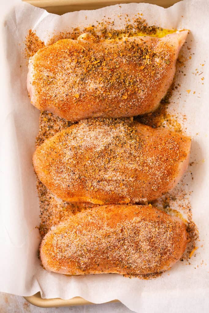 Raw chicken breasts with seasonings on parchment paper.