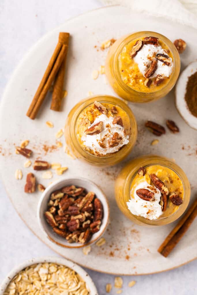 Overhead view of creamy pumpkin pie protein overnight oats served in jars topped with whipped cream and pecans, garnished on a plate with a cinnamon and more pecans.
