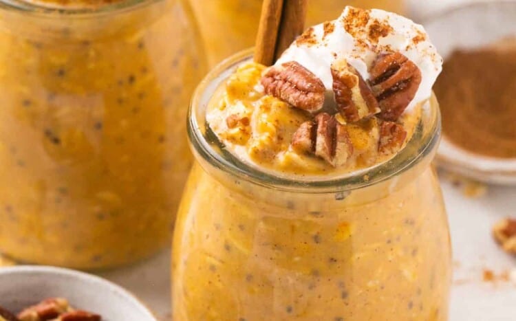 Creamy pumpkin pie protein overnight oats served in jars topped with whipped cream and pecans, garnished with a cinnamon stick.