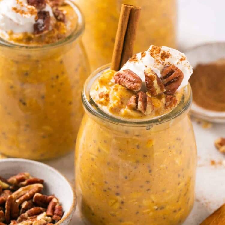Creamy pumpkin pie protein overnight oats served in jars topped with whipped cream and pecans, garnished with a cinnamon stick.