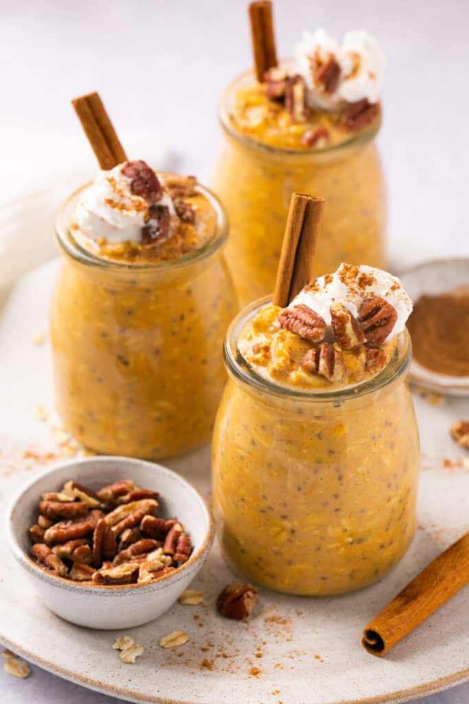 Pumpkin pie protein overnight oats served in jars topped with whipped cream and pecans, garnished with a cinnamon stick.
