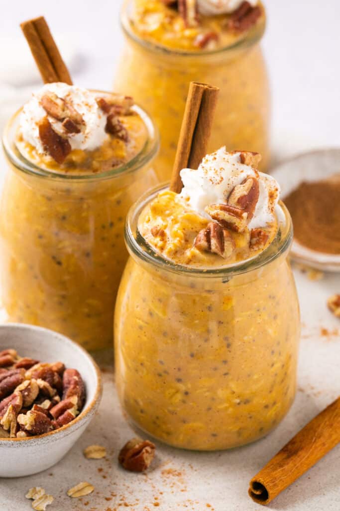 Pumpkin pie protein overnight oats served in jars topped with whipped cream and pecans, garnished with a cinnamon stick.