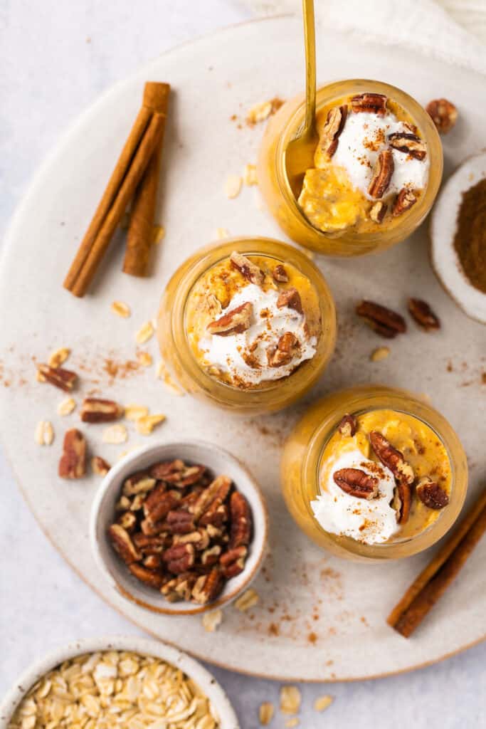 Overhead view of creamy pumpkin pie protein overnight oats served in jars topped with whipped cream and pecans, garnished with a cinnamon stick.