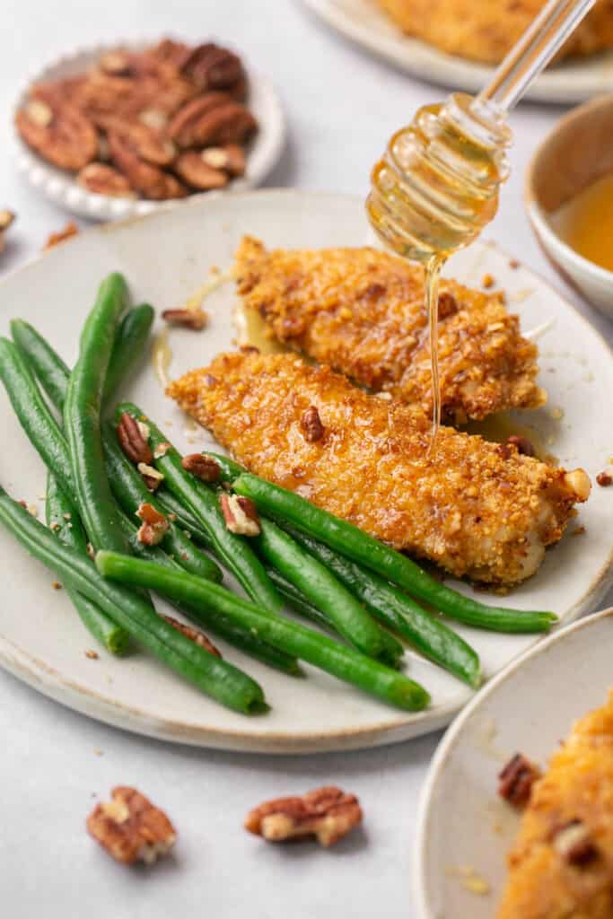 Pecan crusted chicken tenders being drizzled with honey and served on a plate with green beans