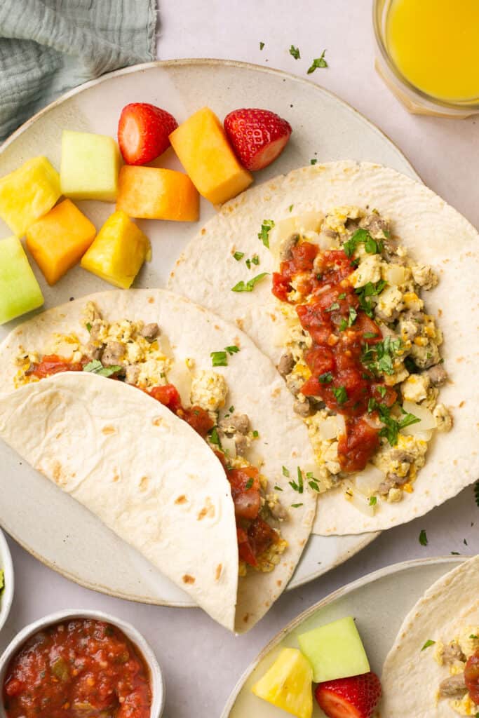 Sausage and cheese breakfast tacos open faced on a plate with fruit