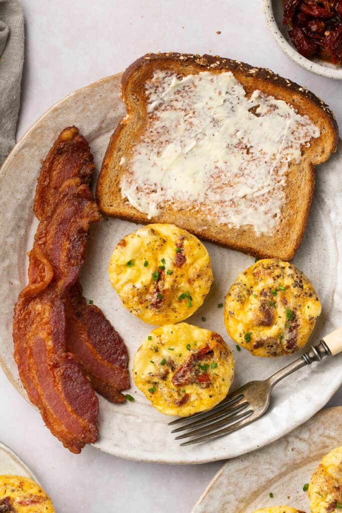 Sun dried tomatoe egg bites, bacon, and toast with butter on a plate.
