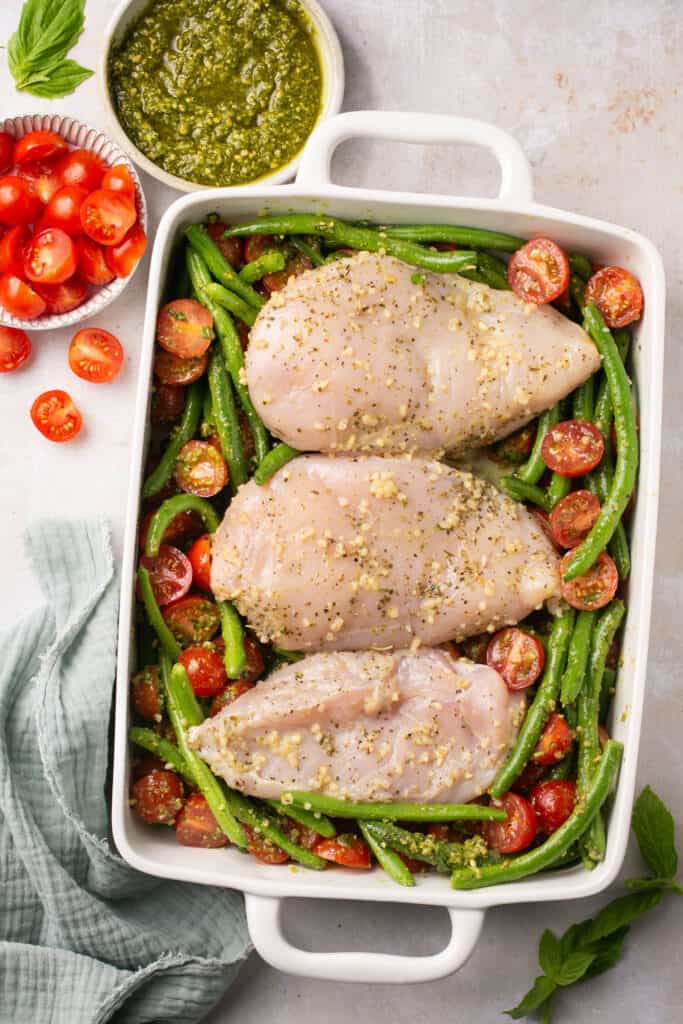Raw chicken breasts with pesto sauce on top of green beans and tomatoes.