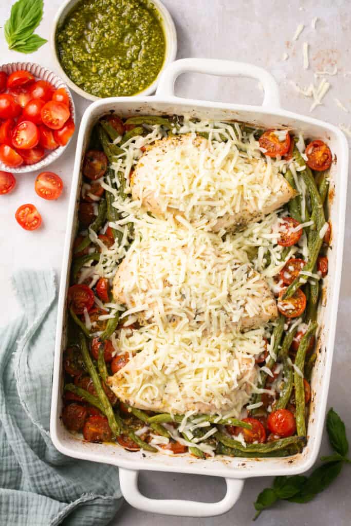 Baked pesto chicken topped with shredded mozzarella cheese in a baking dish.