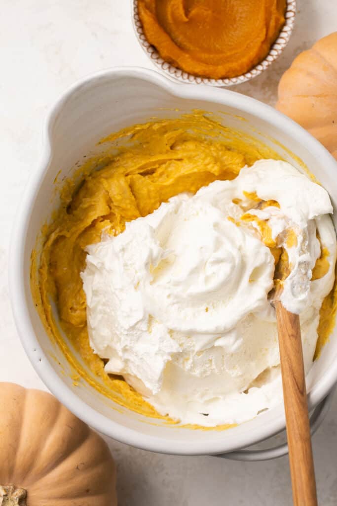 Cool whip being folded into the pumpkin mixture in a mixing bowl with a wooden spoon.