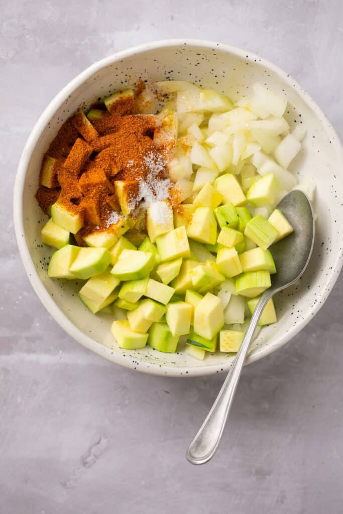 Chopped zucchini and onions and spices in a small bowl with a spoon.