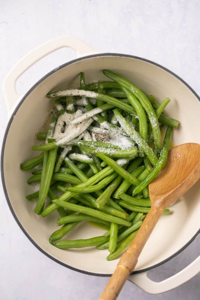 Green beans topped with seasoning in dutch oven with a wooden spoon.