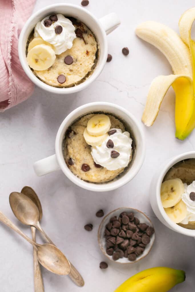 Several banana mug cakes in mugs topped with chocolate chips, bananas, and whipped cream.