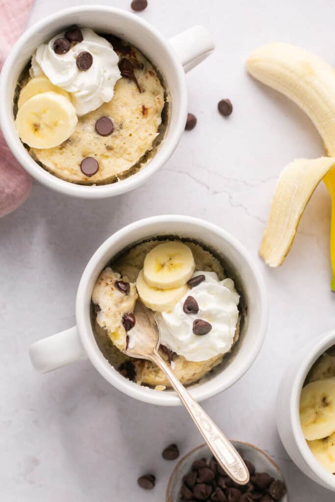 Healthy banana mug cake in a mug with a spoon topped with chocolate chips, bananas, and whipped cream.