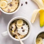 Healthy banana mug cake in a mug with a spoon topped with chocolate chips, bananas, and whipped cream.