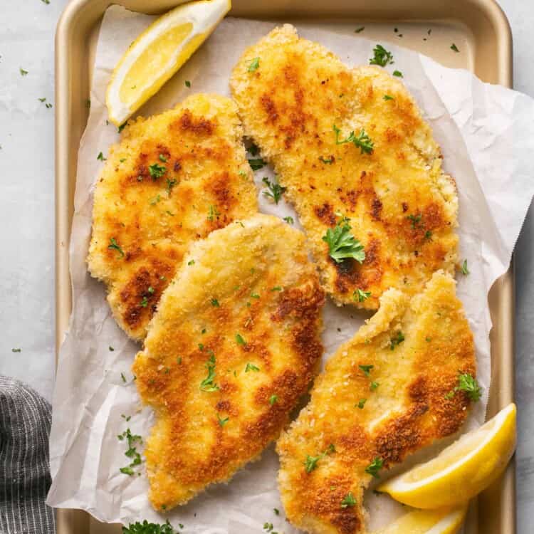 Crispy chicken schnitzel cut into pieces on a tray with parchment paper garnished with lemon slices.