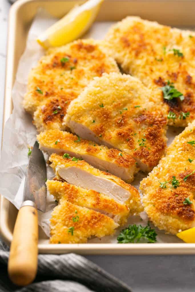 Crispy chicken schnitzel cut into pieces on a tray with parchment paper garnished with lemon slices