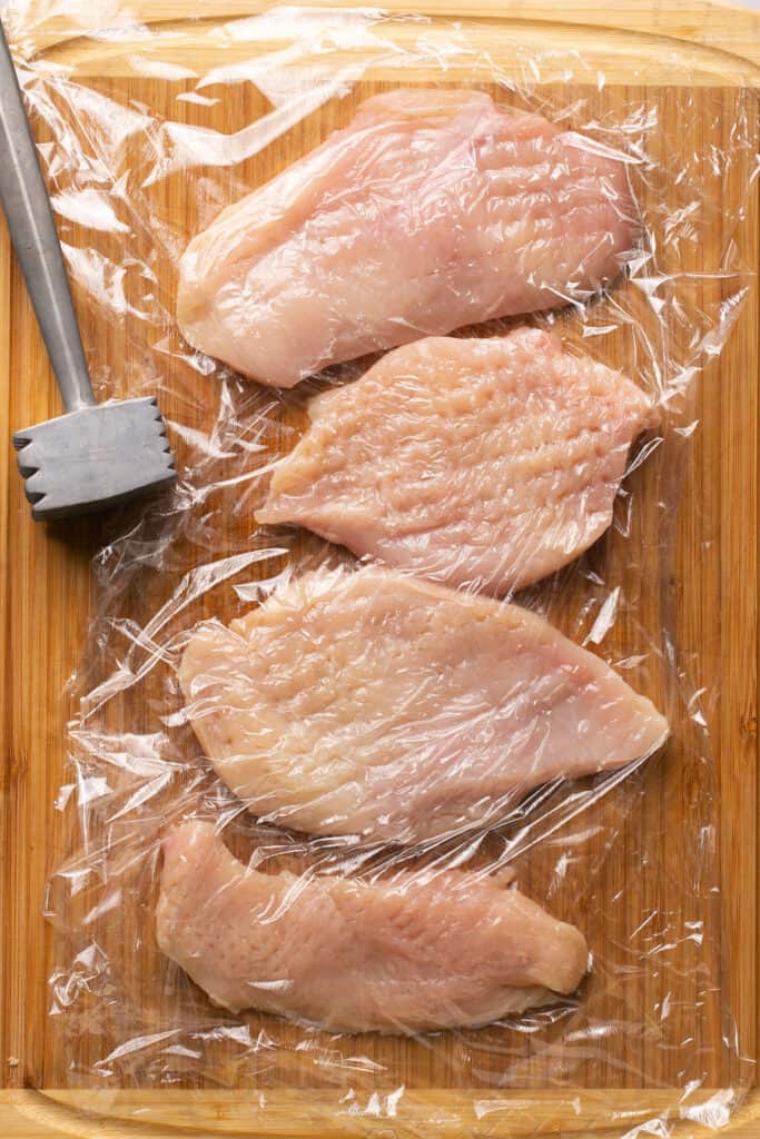 Raw chicken breasts on a cutting board covered with plastic wrap.