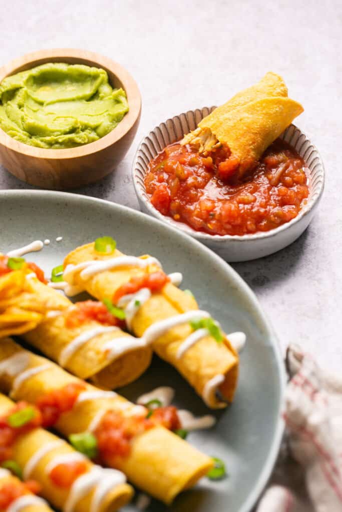 Air fryer chicken taquitos drizzled iwth sour cream and salas on a plate, one taquito being dipped in a side of salsa