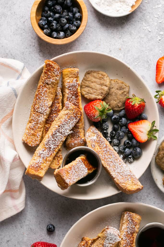 Protein french toast sticks on a plate with fruit, turkey sausage, and side of syrup.