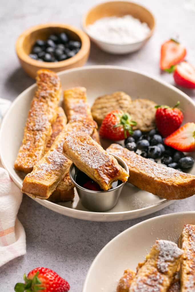 Protein french toast sticks on a plate with fruit, turkey sausage, and side of syrup