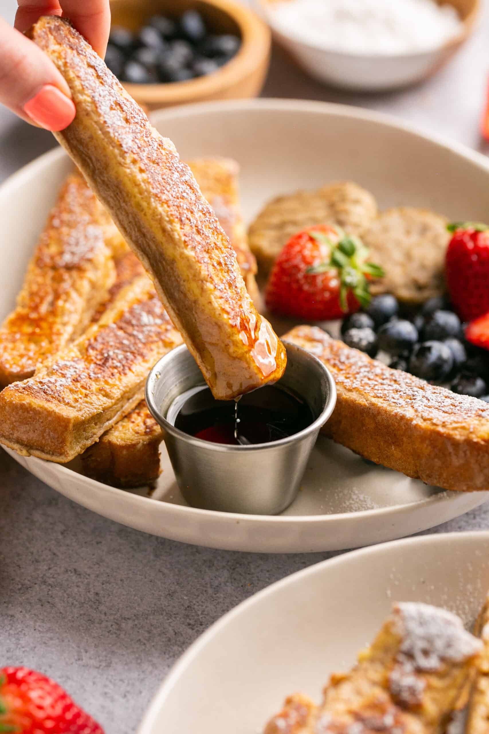 Protein french toast sticks on a plate with fruit, turkey sausage, and side of syrup.