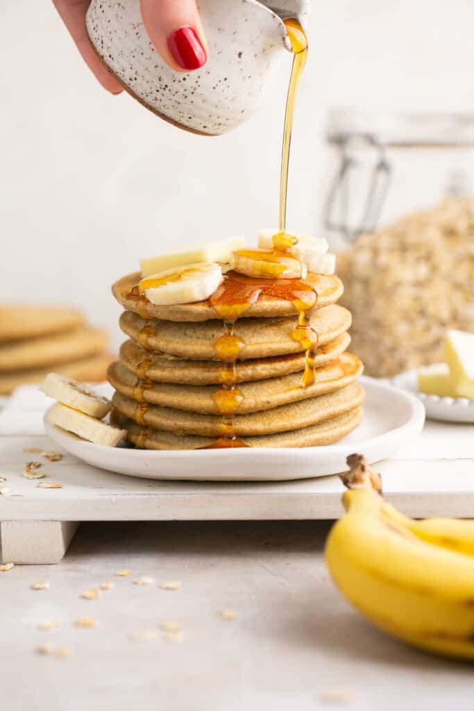 Blender banana oatmeal pancakes stacked up on a plate and topped with banana slices, butter, and maple syrup.